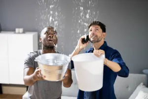 In a dimly lit living room, two men frantically hold buckets to catch water pouring from a leaking ceiling. The first man stands poised with his phone pressed to his ear, exuding concern and urgency as he likely discusses the emergency with a plumber. His companion focuses intently on positioning the bucket perfectly under the leak, ensuring not a drop touches the floor. This vivid scene highlights the immediate chaos of an unexpected plumbing disaster. For residents in Lafayette IN, calling Plumbing Solutions could be their quickest relief amid this watery crisis.
