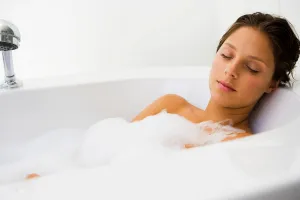 A person unwinds in a luxurious bubble-filled bathtub, eyes gently closed and face framed by a serene smile, embodying pure relaxation. The minimalist design of the bathroom is elegantly highlighted with pristine white walls and sleek fixtures, creating an ambiance of tranquility and homey comfort. Lafayette's expert plumbing ensures every detail contributes to this peaceful oasis.